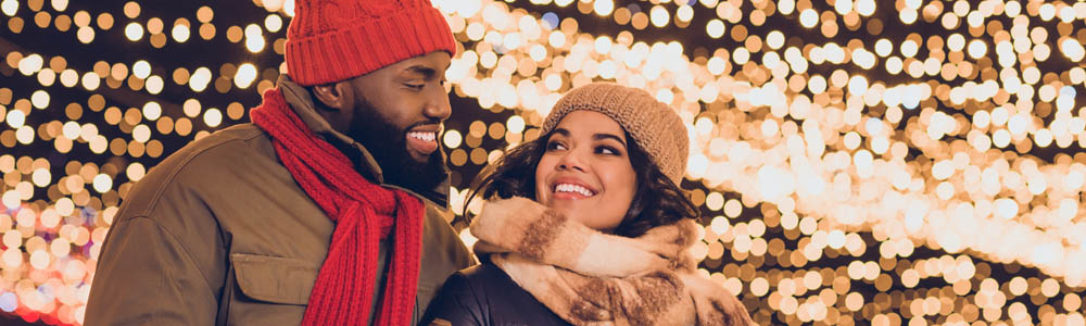 Couple dressed in their winter gear (with hats & scarves), smiling at each other with twinkling Christmas lights in the background Couple dressed in their winter gear (with hats & scarves), smiling at each other with twinkling Christmas lights in the background